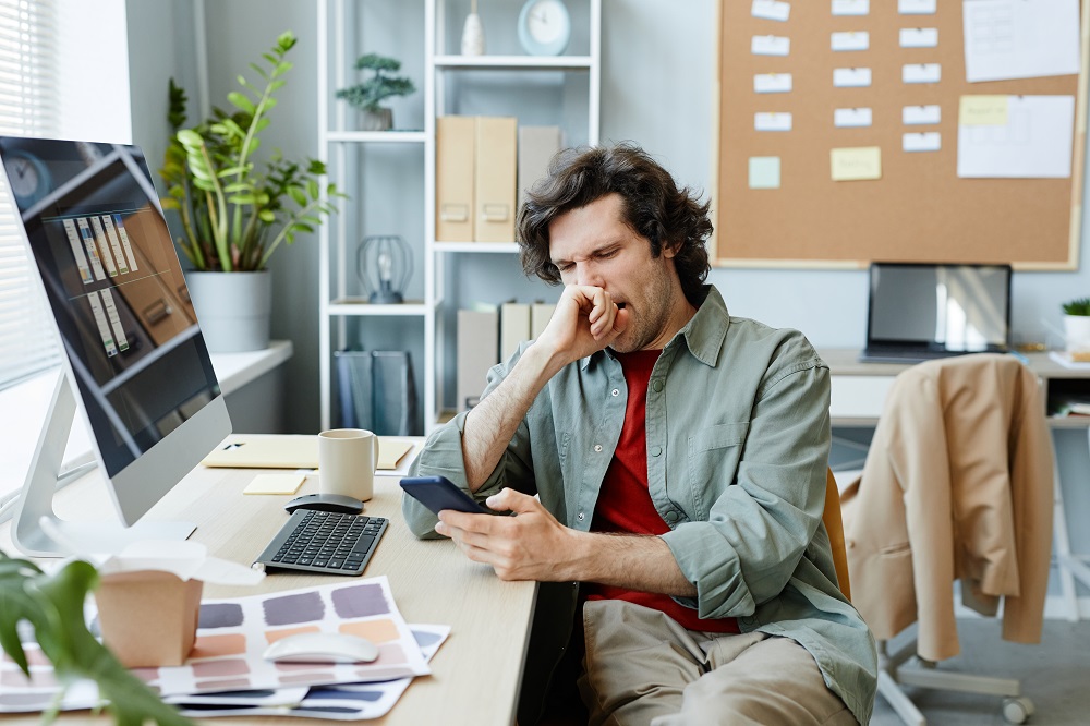 Portrait of Caucasian young man yawning at office workplace and scrolling social media via phone